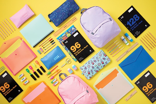 Flat lay of various school supplies including backpacks, notebooks, folders, pens, pencils, and highlighters arranged on a bright yellow background.